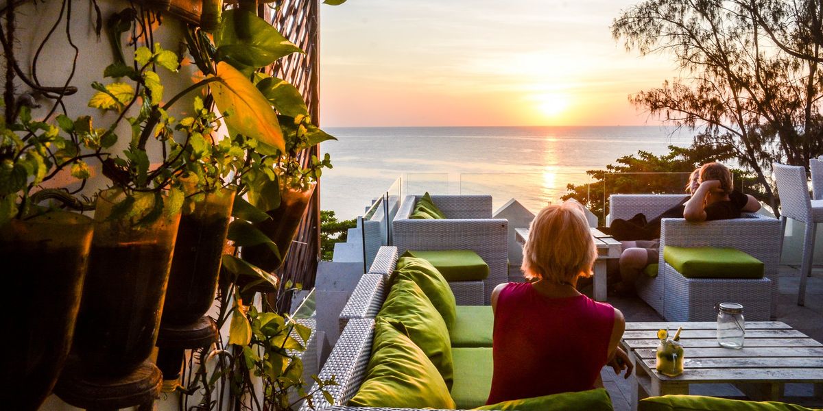 Woman sitting at a table with a cocktail overlooking the ocean at sunset